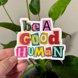 Colorful “Be a Good Human” vinyl sticker with multicolor cutout letters and white border, held in front of green plant leaves indoors.