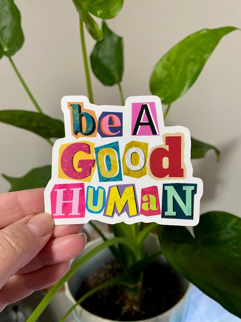 Colorful “Be a Good Human” vinyl sticker with multicolor cutout letters and white border, held in front of green plant leaves indoors.