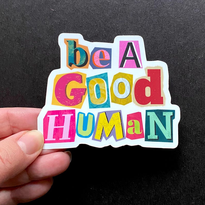 Colorful “Be a Good Human” vinyl sticker with bold cutout letters in pink, yellow, red, and blue, shown held by hand against a black background.