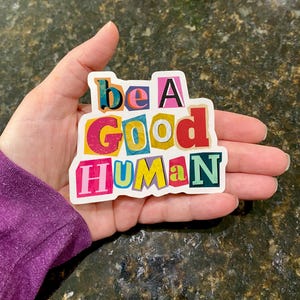 Colorful “Be a Good Human” vinyl sticker with bold multicolor letters and white border, held in a hand outdoors on a textured ground background.