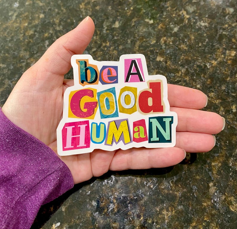 Colorful “Be a Good Human” vinyl sticker with bold multicolor letters and white border, held in a hand outdoors on a textured ground background.