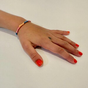 May include: A hand with bright orange nail polish is wearing a colorful friendship bracelet. The bracelet is made of woven string in pink, blue, and yellow.