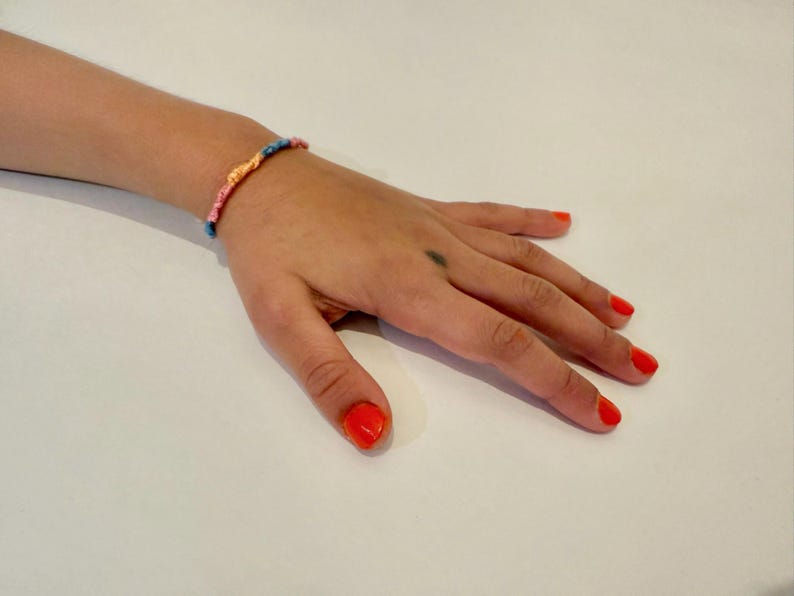 May include: A hand with bright orange nail polish is wearing a colorful friendship bracelet. The bracelet is made of woven string in pink, blue, and yellow.