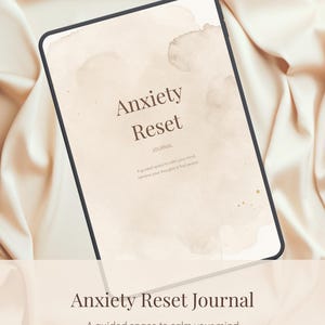 Op de afbeelding: Een digitale tablet met een journal-omslag getiteld Anxiety Reset met een beige aquarelachtergrond. De tekst luidt Anxiety Reset JOURNAL A guided space to calm your mind, reframe your thoughts & find peace. Het scherm heeft een zwarte rand.