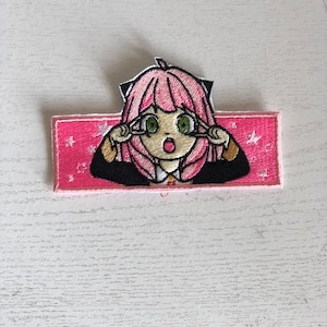 May include: Embroidered iron-on patch featuring a pink-haired anime character with green eyes and a surprised expression. The character is wearing a black and gold outfit and has cat ears. The patch is set against a pink background with white stars.