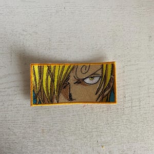May include: A yellow and brown embroidered patch featuring a close-up of an anime character's eye with a yellow iris and a black eyebrow.