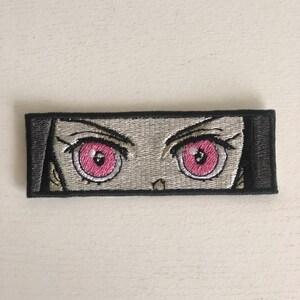 May include: Embroidered patch featuring a close-up of a pair of anime eyes with pink irises and black pupils. The eyes are set against a white background and framed by black outlines.
