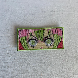 May include: Embroidered patch featuring a stylized anime character's eyes. The patch has a rectangular shape with a green border. The eyes are green with black outlines, and the hair is pink and green.