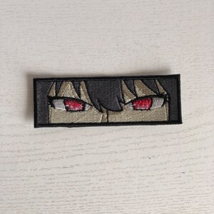May include: Embroidered patch featuring anime-style eyes with red irises and dark hair. The patch is rectangular with a black border and is set against a light-colored wooden surface. The design is detailed and appears to be machine-embroidered.