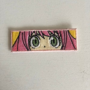 May include: Embroidered patch featuring a cartoon anime girl with pink hair and green eyes. The patch has a yellow background and a pink border.