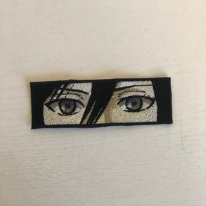 May include: Embroidered patch featuring a pair of anime-style eyes. The eyes are detailed with shades of gray and beige, set against a black rectangular background. The patch is designed for application on clothing or accessories.