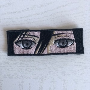 May include: Embroidered patch with a pair of eyes looking directly at the viewer. The eyes are gray with black pupils and are surrounded by pink skin. The patch is black with white stitching.