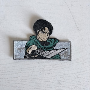 May include: Embroidered patch featuring a character from the anime Attack on Titan. The character is depicted with black hair, a green jacket, and a sword. The patch is rectangular and has a white background.