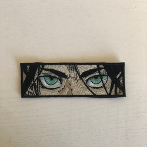 May include: Black and white embroidered patch with a close-up of a person's eyes. The eyes are blue and the person's face is partially visible.