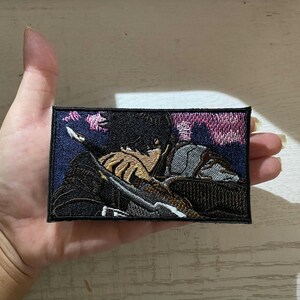 May include: Embroidered patch featuring a detailed anime-style illustration of a person with black hair, holding a weapon. The patch has a rectangular shape with a dark blue border and a background with pink and purple hues.