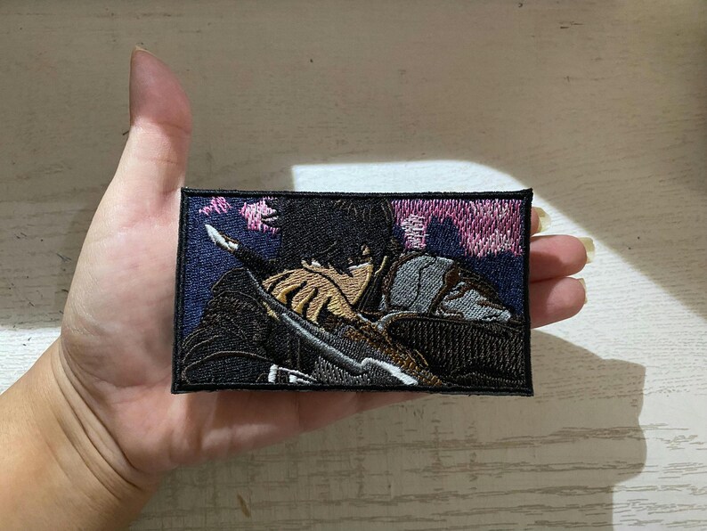 May include: Embroidered patch featuring a detailed anime-style illustration of a person with black hair, holding a weapon. The patch has a rectangular shape with a dark blue border and a background with pink and purple hues.