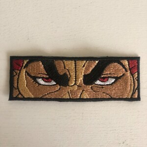May include: Embroidered patch featuring a close-up of a character's face with angry red eyes. The character's face is brown and the background is black.