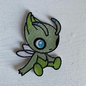 May include: A green and white embroidered patch featuring a cartoon-style creature with large blue eyes, small wings, and a smiling expression. The patch has a black border and is likely designed for application on clothing or accessories.