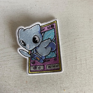 May include: Embroidered patch of a blue and white cartoon creature with large eyes, resembling a Pokemon card. The card has a purple background with the text "EW 30" and "RETREAT" in yellow.