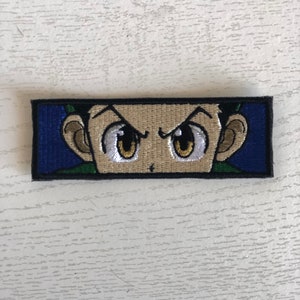 May include: Embroidered patch featuring a cartoon character with green hair, brown eyes, and a serious expression. The character's eyes are wide open and the eyebrows are furrowed. The patch is rectangular and has a black border.