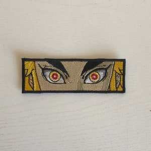 May include: Embroidered patch with a close-up of a pair of eyes with red pupils and yellow irises. The eyes are surrounded by a gold background and a black border.