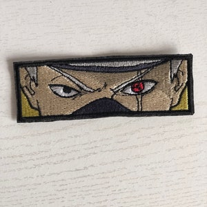 May include: Embroidered patch featuring a close-up of a character's face with a single red eye. The character has white hair and a dark headband. The patch is surrounded by a black border.