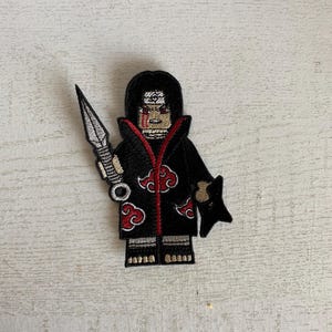 May include: Embroidered patch featuring a stylized character in a black cloak with red cloud designs. The character holds a silver kunai and a black shuriken. The patch has a red zipper and is on a white background.