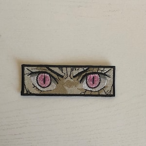 May include: Embroidered patch featuring a pair of stylized eyes with pink irises and detailed white and tan stitching. The patch has a black border and a rectangular shape, suitable for application on clothing or accessories.