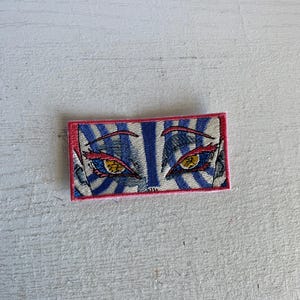 May include: Embroidered patch featuring a stylized eye design. The patch has a rectangular shape with a pink border. The eyes are yellow with red accents, set against a blue and white striped background.