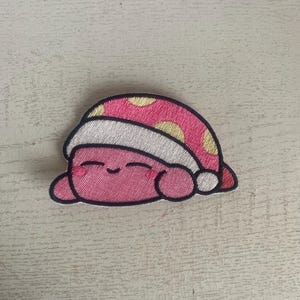 May include: Pink and yellow embroidered patch of a sleeping cartoon character wearing a white and yellow hat.