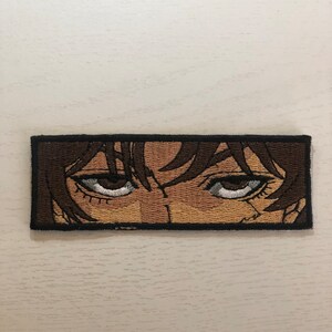 May include: Embroidered patch featuring a close-up of a person's eyes with brown hair. The eyes are wide open and staring directly at the viewer.
