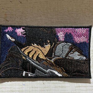 May include: Embroidered patch featuring a detailed illustration of a figure in a dark outfit, holding a sword. The design includes shades of black, brown, gray, and pink, set against a dark blue background. The patch has a black border.