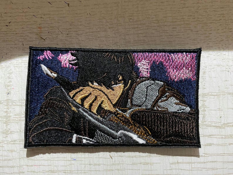 May include: Embroidered patch featuring a detailed illustration of a figure in a dark outfit, holding a sword. The design includes shades of black, brown, gray, and pink, set against a dark blue background. The patch has a black border.