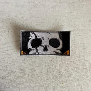 May include: A black and white embroidered patch featuring a skull with a cracked bone and one eye visible. The skull is surrounded by a yellow border.