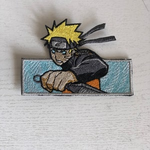 May include: Embroidered patch featuring a yellow-haired anime character with blue eyes, wearing a headband with a swirl symbol, and a black and gray outfit. The character is holding a weapon and is set against a blue background.