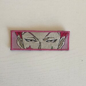May include: Embroidered patch featuring a stylized face with pink hair and a pink border. The eyes are detailed with a focused expression. The patch is rectangular and likely designed for application on clothing or accessories.
