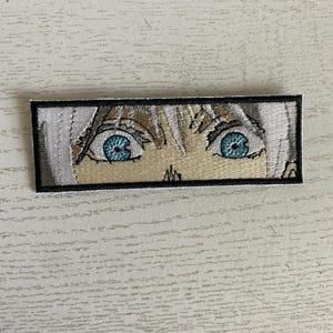 May include: Close-up of a white embroidered patch with two blue eyes. The eyes are looking directly at the viewer.