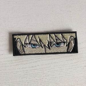 May include: Embroidered patch featuring anime-style eyes with light blonde hair and striking blue eyes. The patch is rectangular with a black border. The design is detailed, with visible eyelashes and shading, creating a captivating look.