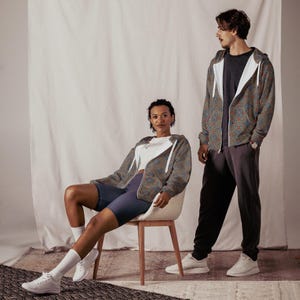 May include: Two people wearing matching patterned zip-up hoodies. The person on the left is sitting on a chair and wearing blue shorts. The person on the right is standing and wearing grey pants.