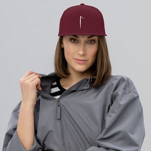 May include: A woman wearing a burgundy baseball cap with a white graphic on the front. The cap has a curved brim and a snap closure. She is also wearing a gray jacket with a zipper.