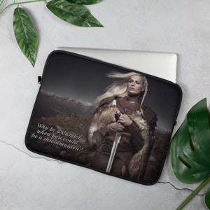 May include: A laptop sleeve featuring a woman in armor holding a sword. The background is a dark, stormy sky with mountains in the distance. The text on the sleeve reads: "Why be a damsel when you could be a shieldmaiden?"