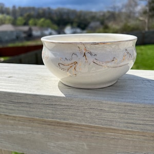 May include: A white ceramic bowl with gold trim and a floral design. The bowl is sitting on a wooden surface.