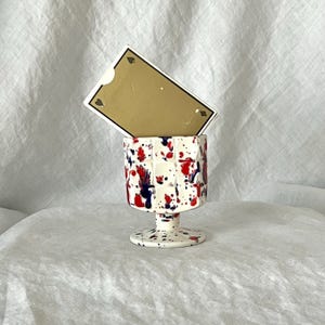 May include: A white ceramic goblet with a speckled design of red and blue paint splatters. A gold playing card is placed inside the goblet. The goblet has a pedestal base and a square-like shape.