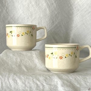 May include: Two cream-colored ceramic mugs with a floral design. Each mug has a handle and a green band at the top. The floral pattern includes yellow, red, and green elements. The mugs are set against a white background.