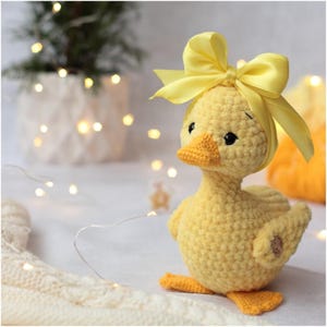 Peut inclure: Un canard jaune en crochet fait &agrave; la main, orn&eacute; d'un n&oelig;ud en ruban jaune sur la t&ecirc;te. Il a un bec jaune, des yeux noirs et des pieds orange. L'arri&egrave;re-plan est flou avec un &eacute;clairage doux.