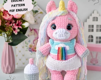 May include: A pink crocheted unicorn amigurumi toy with a rainbow and cloud design on its shirt. The unicorn has a long, colorful mane and a yellow horn. The toy is holding a small, gray crocheted bottle.