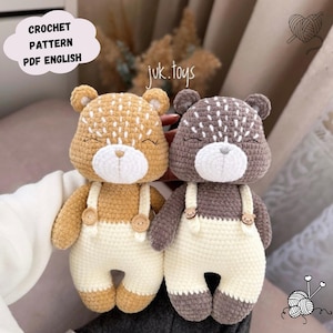 May include: Two crocheted teddy bear toys wearing white overalls with brown buttons. The teddy bears have brown and gray fur and white faces with embroidered eyes and noses. The bears are sitting on a white surface.