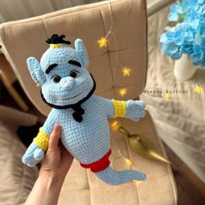 May include: A light blue crocheted plush toy shaped like a genie with a black beard, yellow wrist cuffs, and a red waistband. The figure has large eyes, pointed ears, and a small topknot. The text @teddy_knitted is visible on the right side.