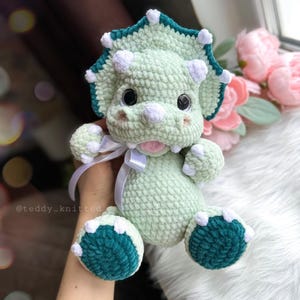 May include: A green crocheted triceratops stuffed animal with white accents and a teal frill. The toy has large, black eyes and a pink tongue sticking out.