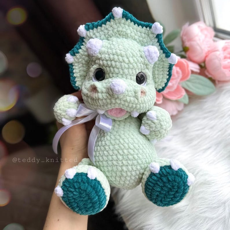 May include: A green crocheted triceratops stuffed animal with white accents and a teal frill. The toy has large, black eyes and a pink tongue sticking out.